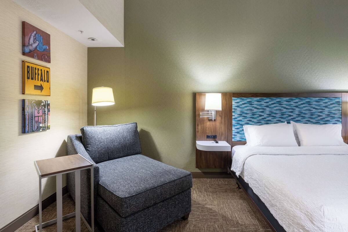 Une chambre d’hôtel avec un fauteuil à revêtement gris, une petite table, une lampe de chevet, un lit blanc avec une tête de lit à motifs bleus et un lavabo fixé au mur.
