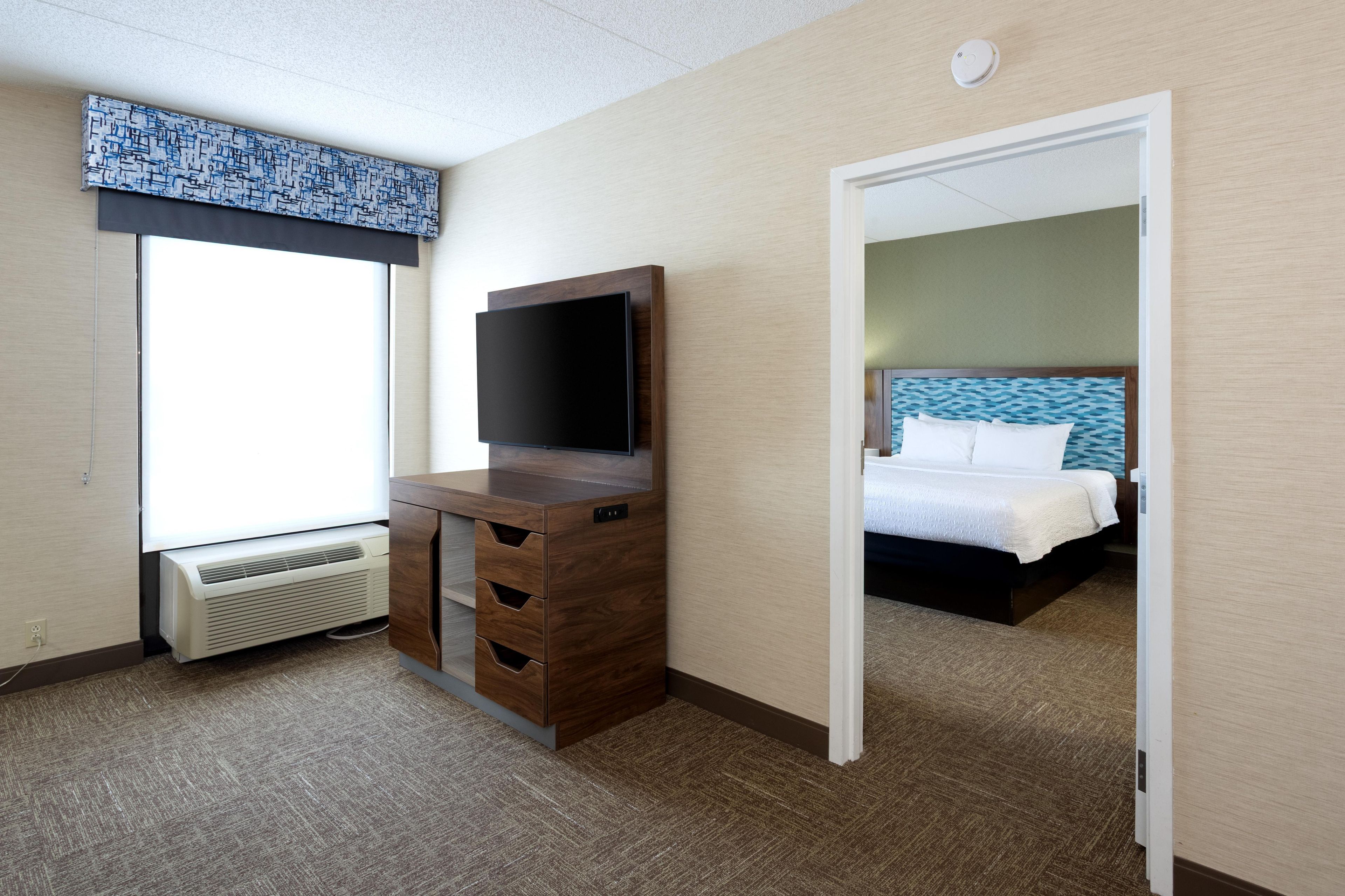 Quarto de hotel com escrivaninha, televisão de tela plana, cama com cabeceira azul e janela com persianas.