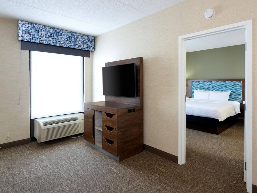 Quarto de hotel com escrivaninha, televisão de tela plana, cama com cabeceira azul e janela com persianas.
