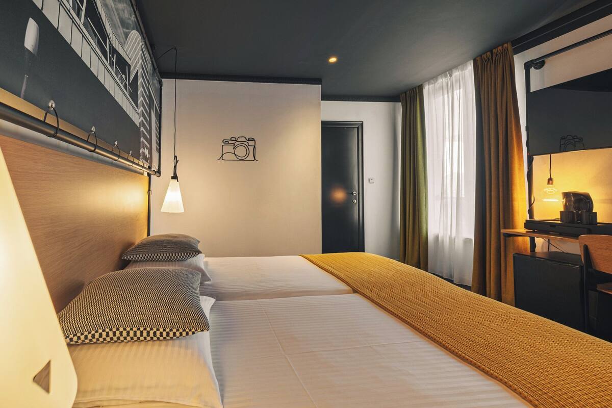 Quarto de hotel com cama, escrivaninha e cadeira. Há uma porta e uma janela com cortinas.