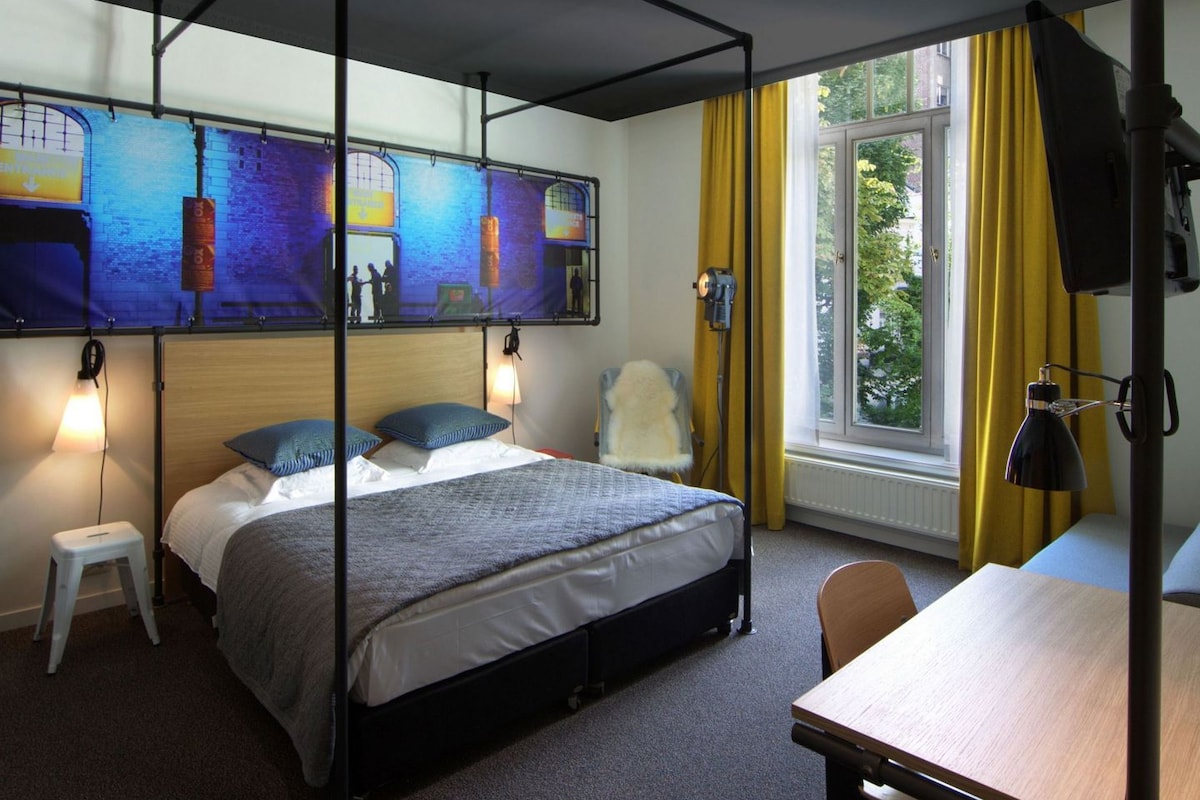 Una cama con dosel con vistas a un mural de paisaje urbano, un taburete pequeño, una mesa y una ventana con cortinas amarillas.