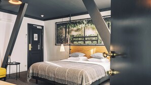 A modern bedroom with a bed, a nightstand, a lamp, and a view of greenery through a window. - Zoom Hotel (Brussels)