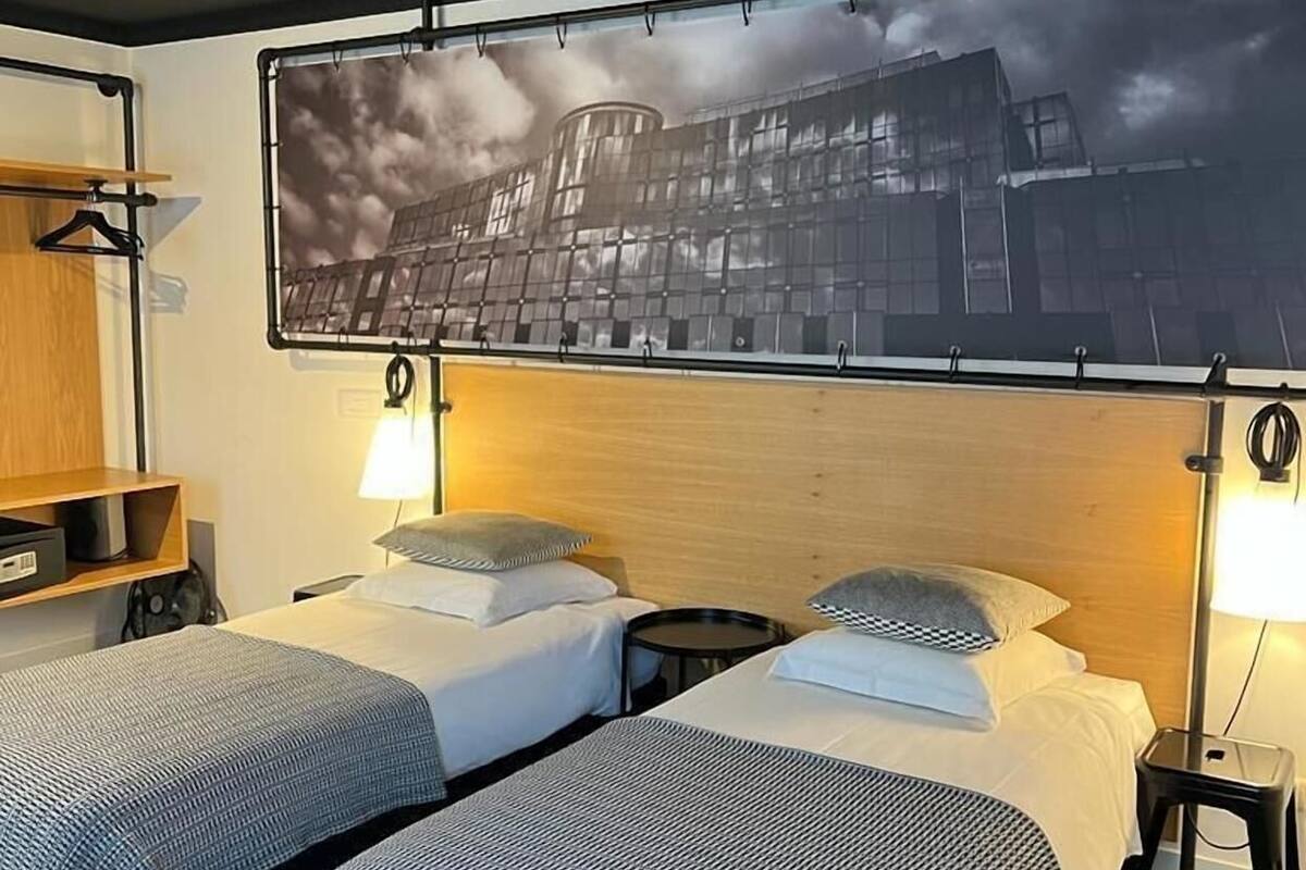 Quarto de hotel com duas camas de solteiro, cabeceira de madeira, televisão presa à parede e uma pequena mesa com abajur.