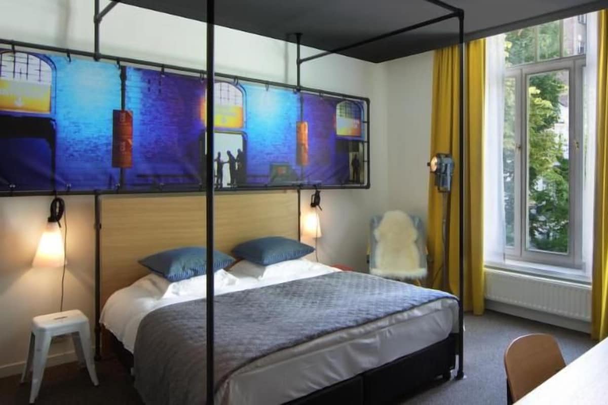 Una habitación de hotel con cama, escritorio, silla, ventana con cortinas y obras de arte en la pared.