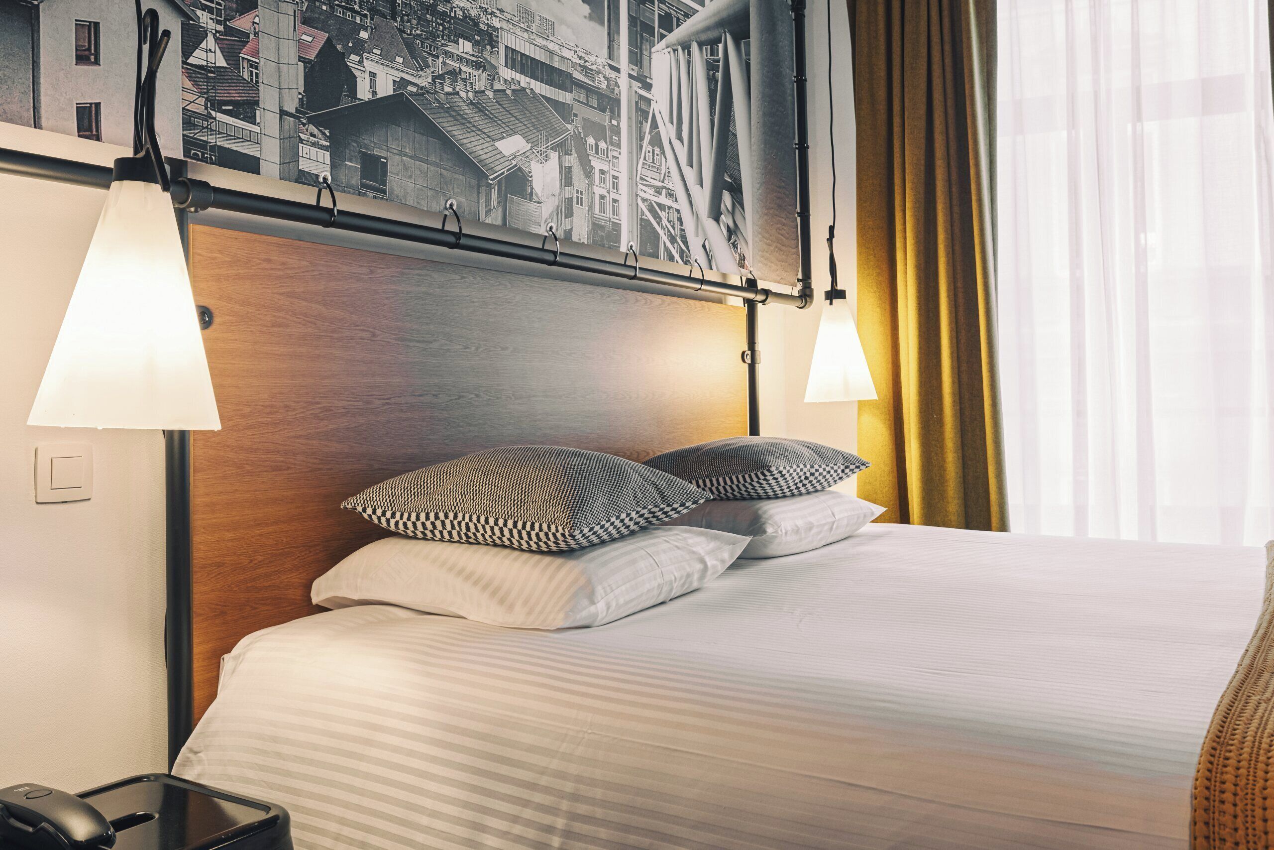A hotel room with a neatly made bed, two bedside lamps, a headboard with a cityscape mural, and a suitcase on the floor.