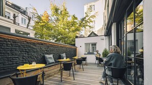 An outdoor seating area with wooden flooring, black and yellow chairs, and a stone wall. - Zoom Hotel (Brussels)