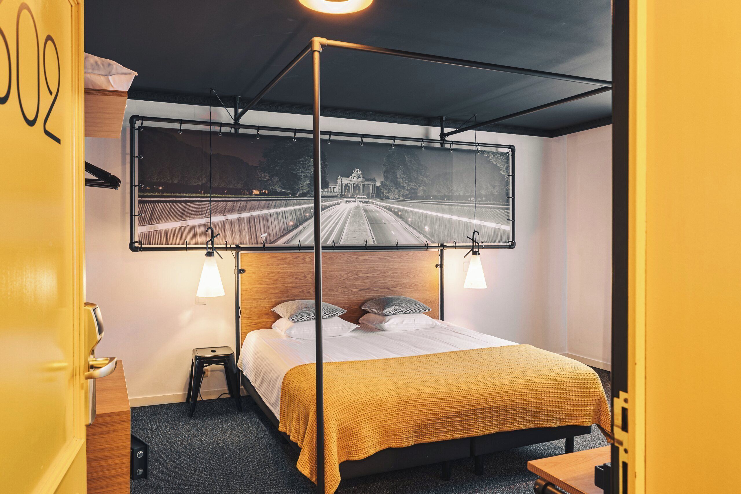 Une chambre d’hôtel avec un lit, une table de chevet, un petit tabouret, une grande fenêtre donnant sur un pont et un téléviseur fixé au mur.