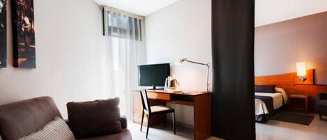 A modern hotel room with a brown sofa, a desk with a computer, a bed, and a large window with sheer curtains.