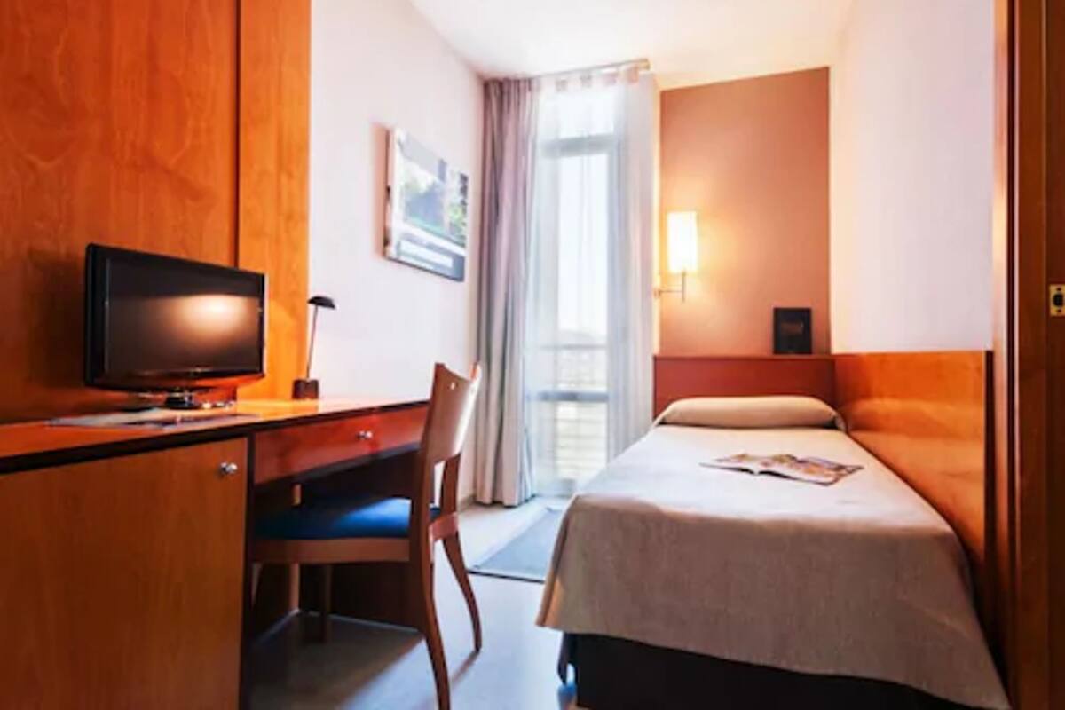 Habitació d'hotel amb llit, escriptori amb televisió, cadira, finestra amb cortines i làmpada penjada a la paret.