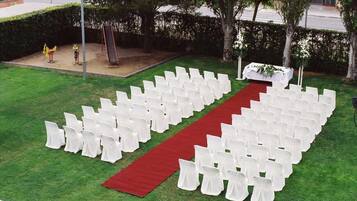 A grassy outdoor area with rows of white chairs arranged in a semi-circle, a red carpet leading to a white table with a floral arrangement.