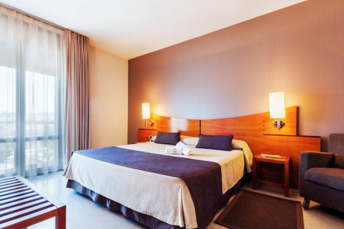 Habitació d'hotel amb un llit gran, dues taules de nit, una cadira, escriptori i vista a l'exterior a través de grans finestres.