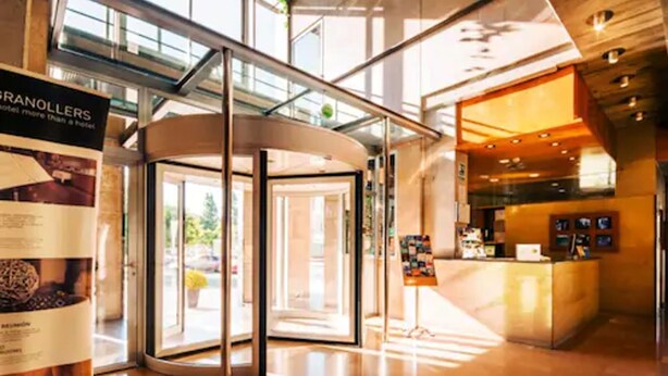 A hotel lobby with a revolving door, a reception desk, and a glass wall offering a view of the outdoors. - Hotel Granollers (Granollers)