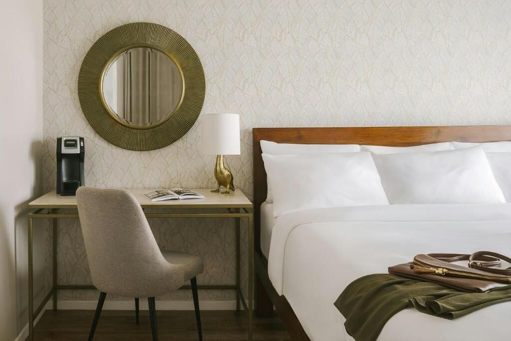 A hotel room with a bed, a desk, a chair, a lamp, a mirror, and a handbag on the bed.