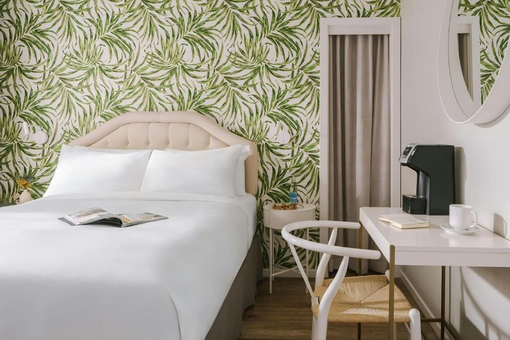 A hotel room with a bed, a small table with a chair, a coffee machine, and a mirror on the wall.