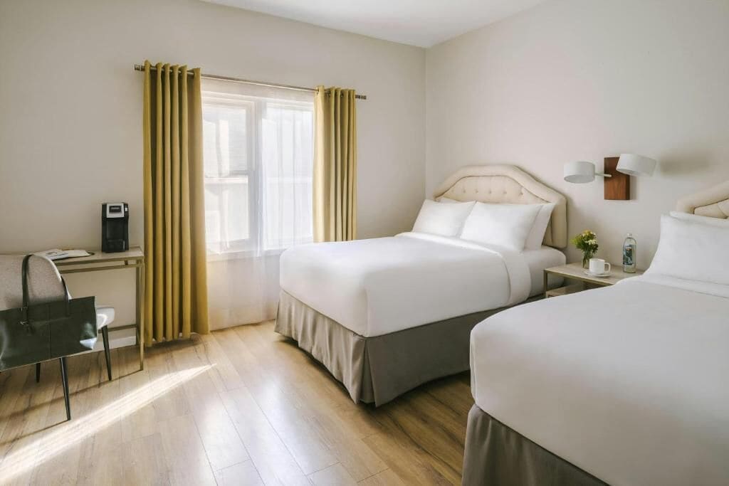 A hotel room with two beds, a desk with a chair, a bedside table with a lamp, and a window with curtains.