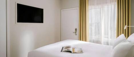 A hotel room with a bed, a television mounted on the wall, and a window with curtains.