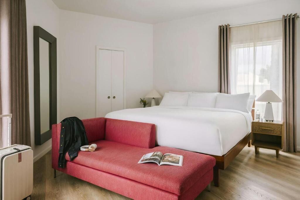 Una habitación de hotel con una cama, un sofá rojo, una mesita de noche con lámpara y una ventana con cortinas.
