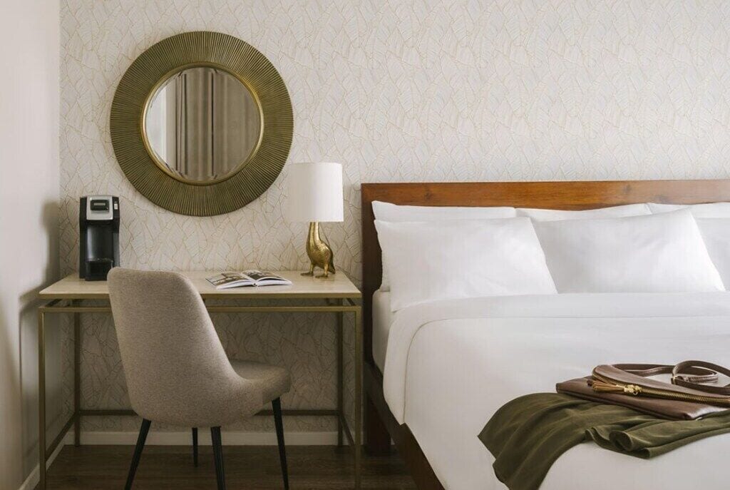 A hotel room with a bed, a desk with a chair, a round mirror, a lamp, and a handbag on the bed.