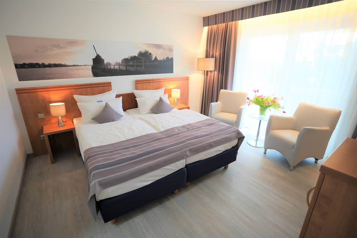 Ein Hotelzimmer mit einem großen Bett, zwei Nachttischen mit Lampen, einem Sessel, einem kleinen Tisch mit Blumen und einem Bild an der Wand.