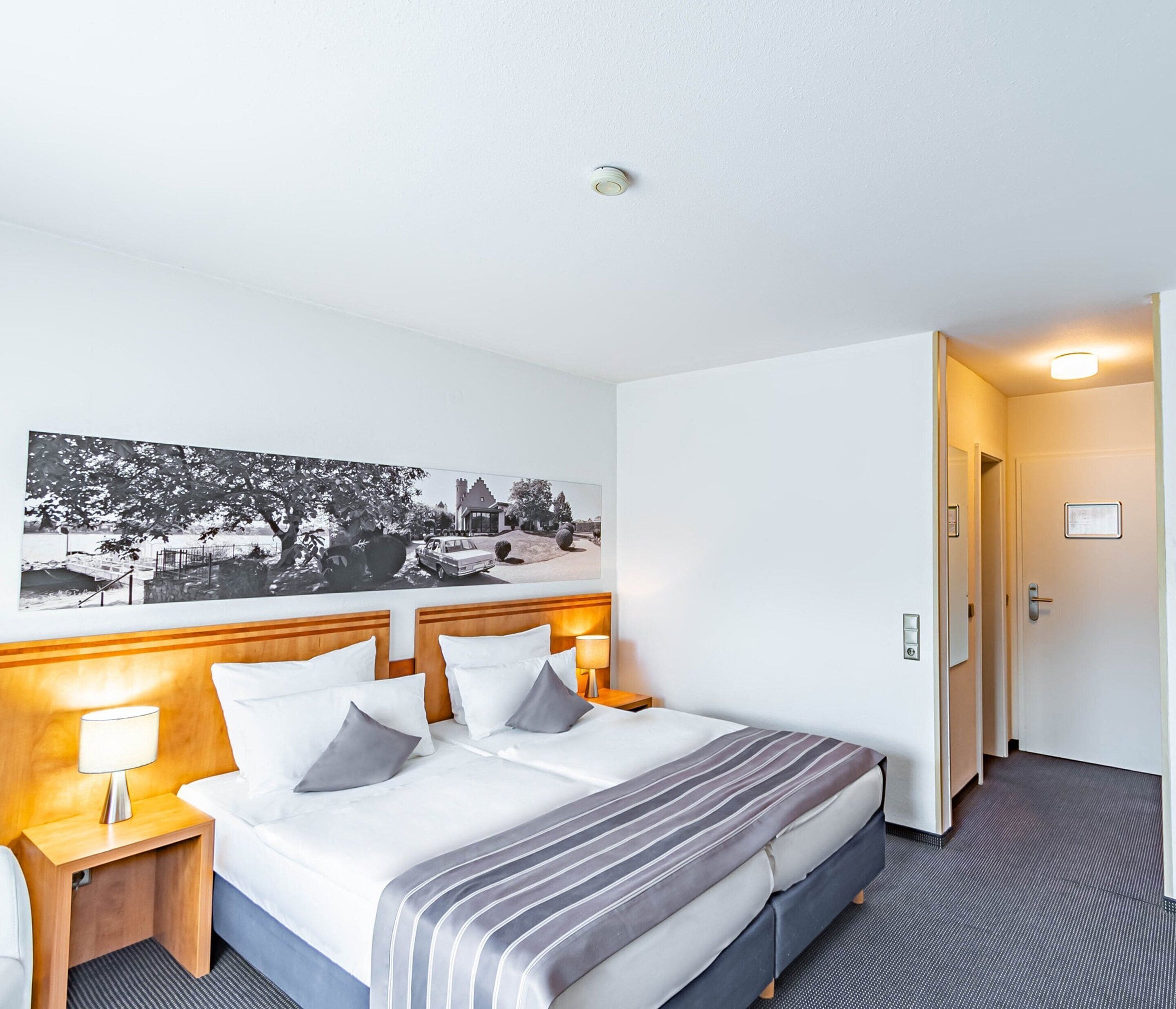 A hotel room with a bed, a nightstand, a lamp, a framed picture on the wall, and a door.
