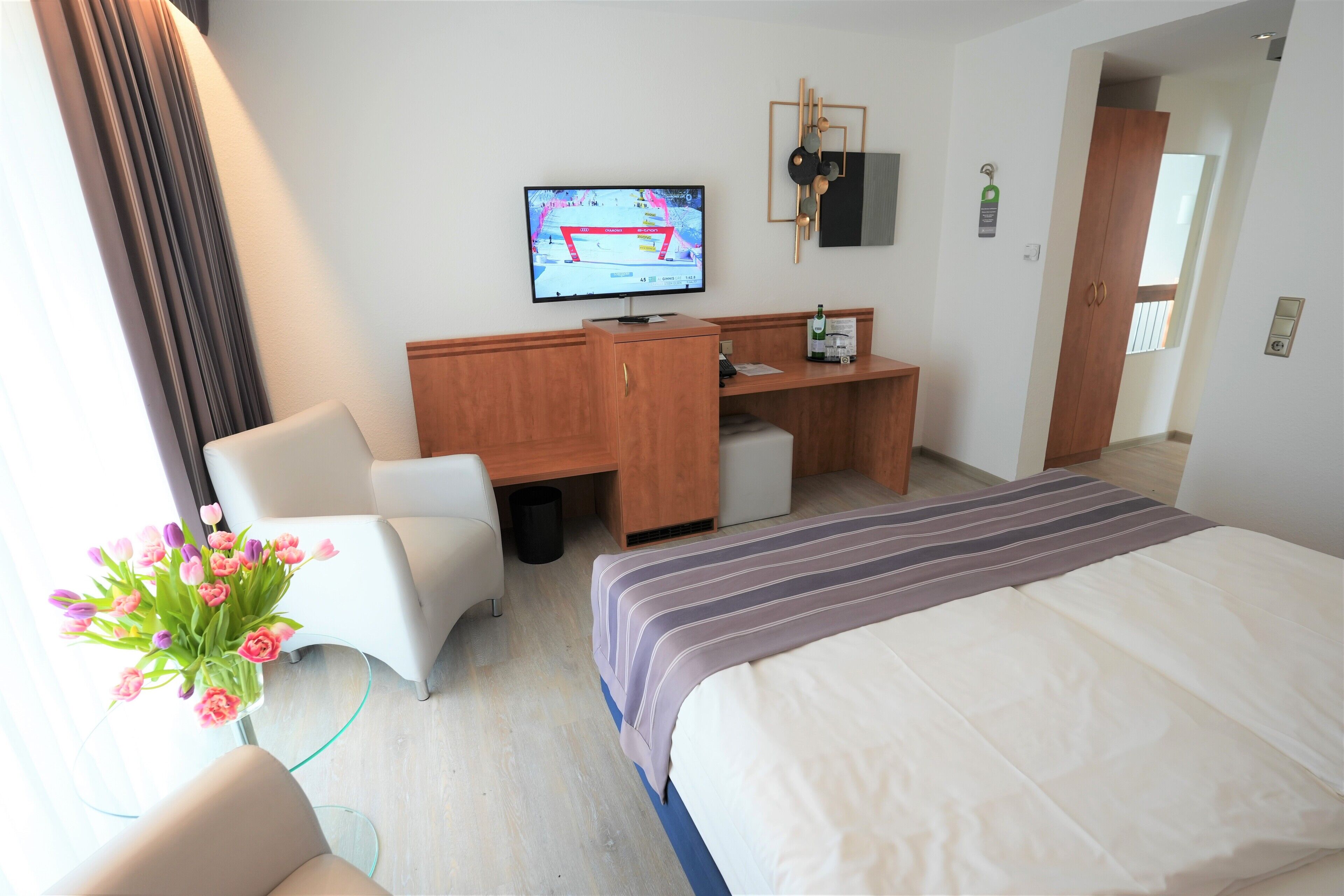 A hotel room with a bed, a desk, a chair, a television, and a vase of flowers.