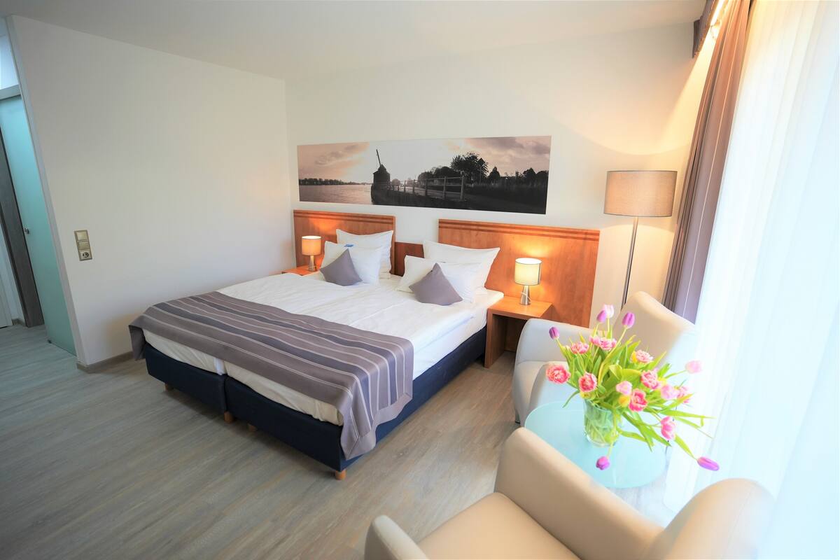 Ein Hotelzimmer mit einem großen Bett, zwei Nachttischen mit Lampen, einem Sessel, einer Vase mit Blumen und einem Bild an der Wand.
