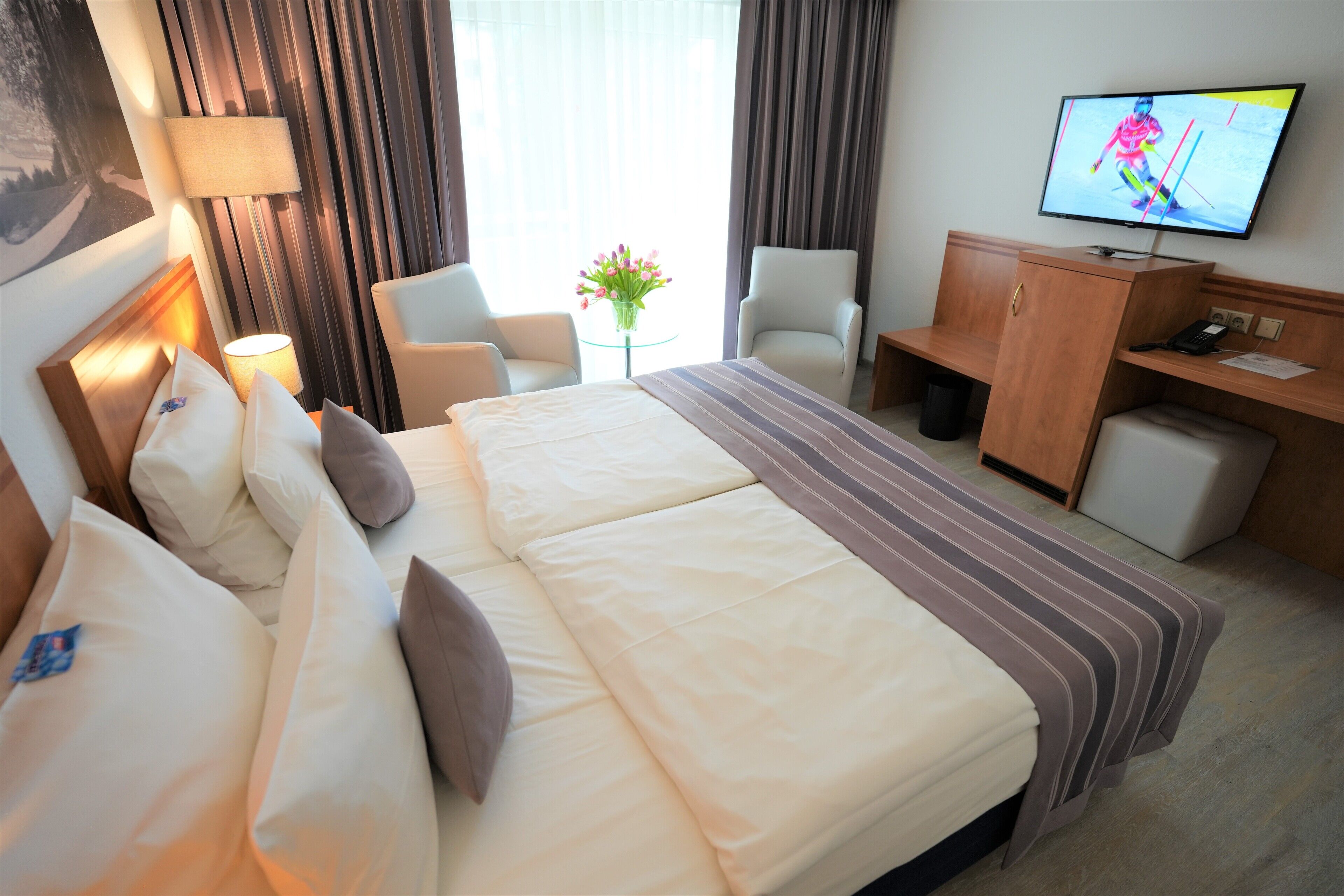 Ein Hotelzimmer mit einem großen Bett, zwei Sesseln, einem Schreibtisch und einem an der Wand befestigten Fernseher.