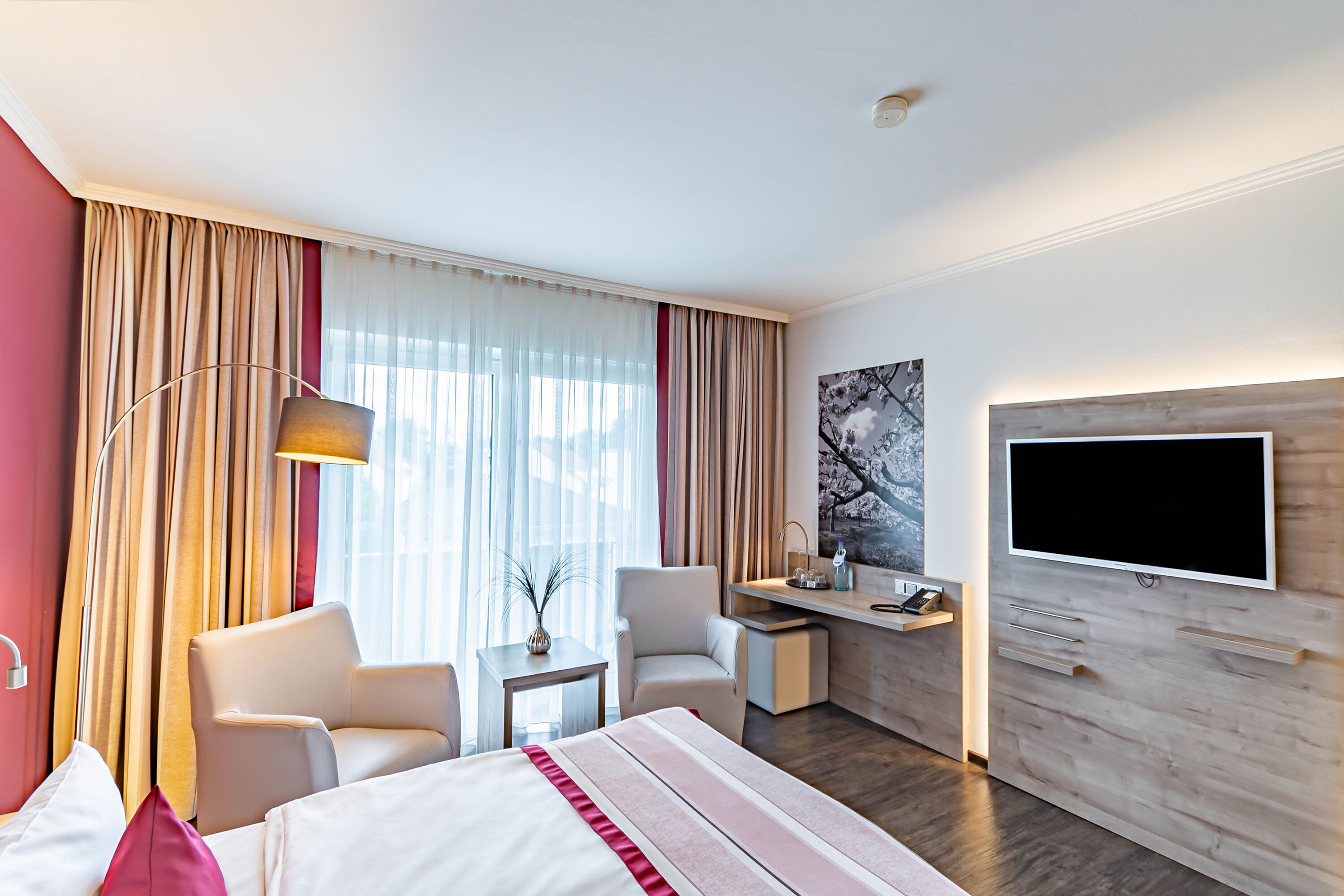 A hotel room with a bed, two chairs, a desk, a TV, and a painting on the wall.