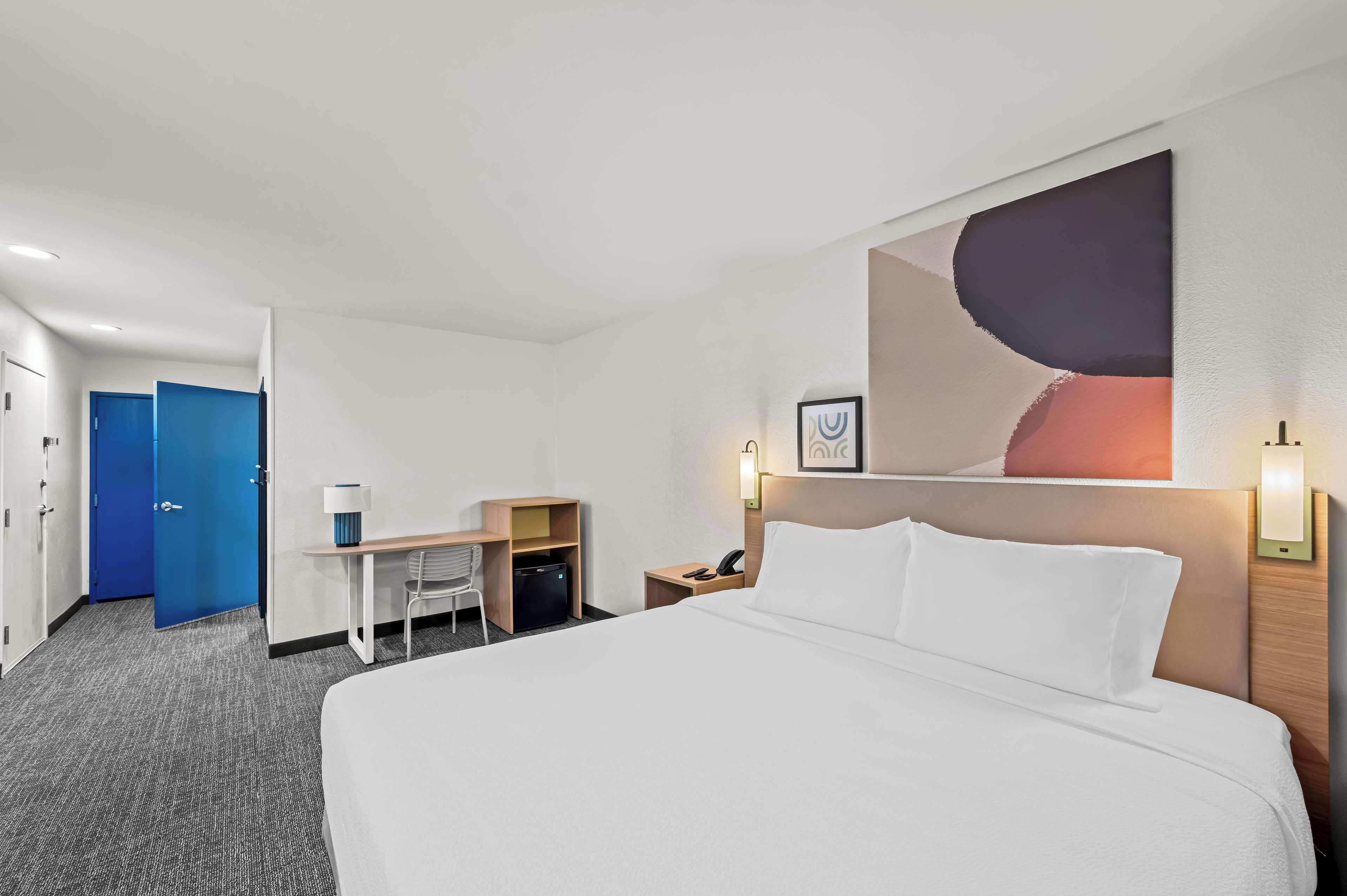 Una habitación de hotel con una cama grande, un escritorio con silla, una pequeña mesa de trabajo con lámpara, un teléfono y un cuadro en la pared.
