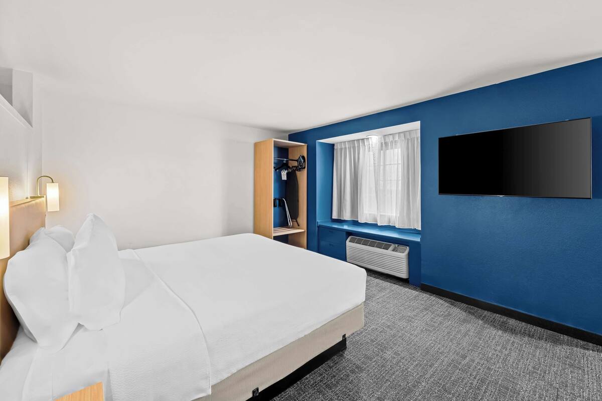 Ein Hotelzimmer mit Bett, Flachbildfernseher an der Wand, Fenster mit Vorhängen und einer fest installierten Heizung.