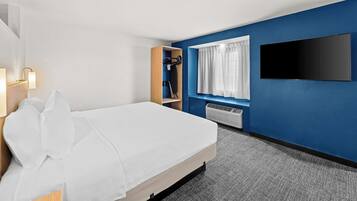 A hotel room with a bed, a flat-screen TV mounted on the wall, a window with curtains, and a built-in heater.