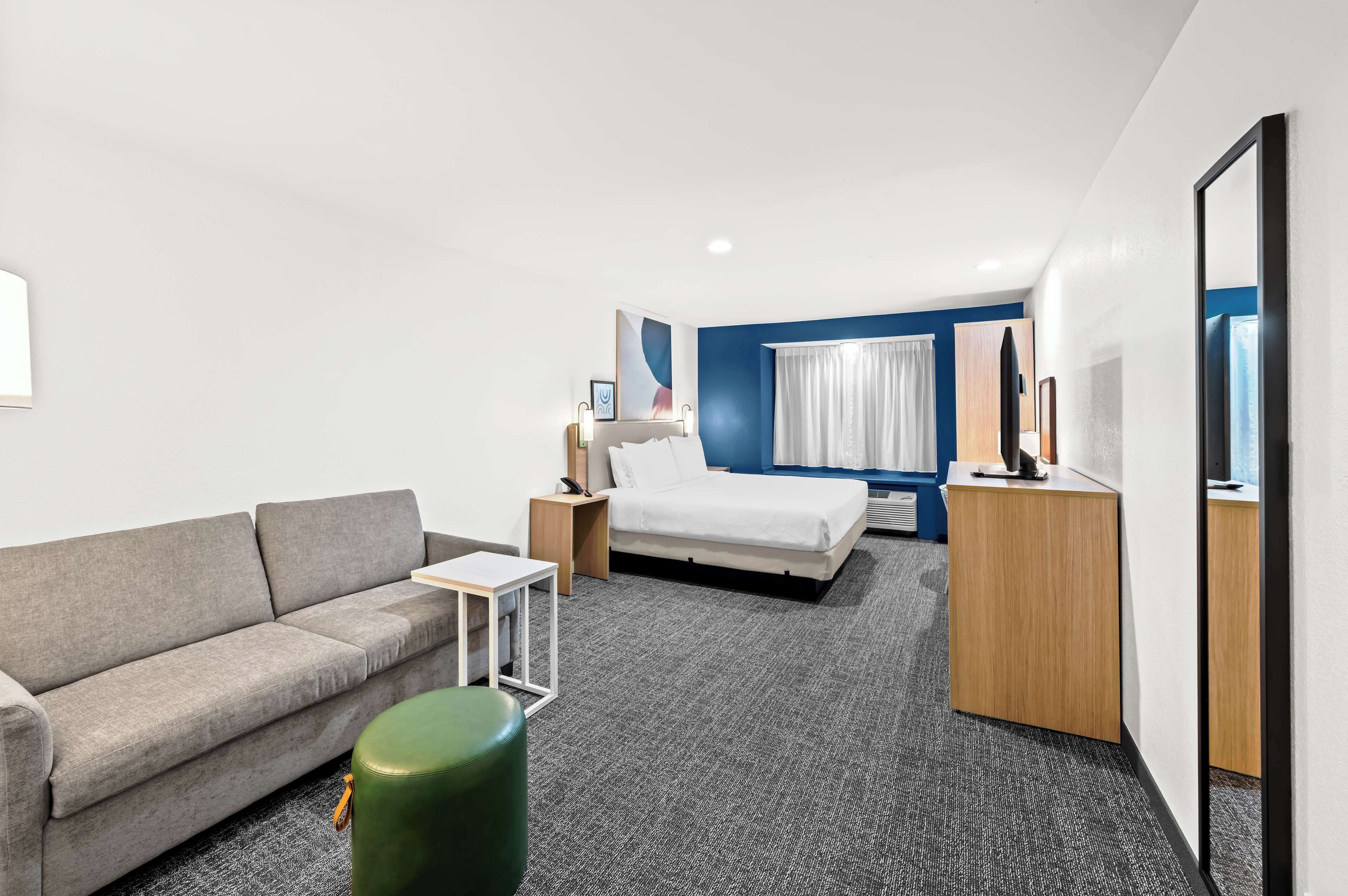 Une chambre d’hôtel moderne avec un canapé gris, un lit simple, une petite table et un pouf vert.