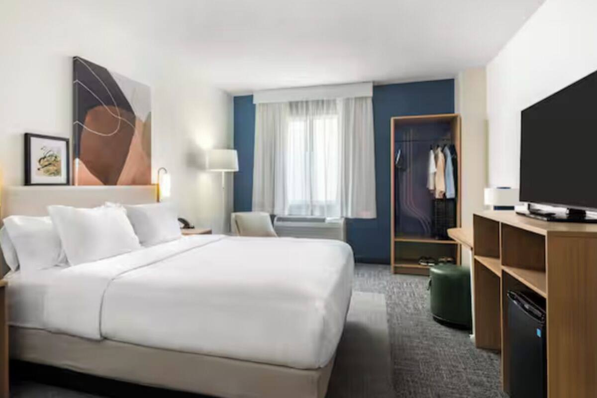 Ein Hotelzimmer mit einem großen Bett, einem Schreibtisch, einem Stuhl, einem Kleiderschrank, einem Fernseher und einem Fenster mit Vorhängen.
