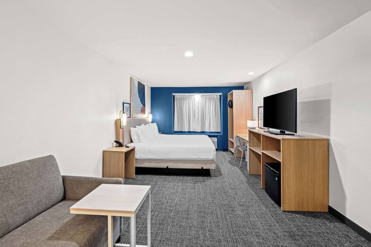 Ein Hotelzimmer mit Bett, Fernseher, Schreibtisch, Sofa und einem Fenster mit Vorhängen.