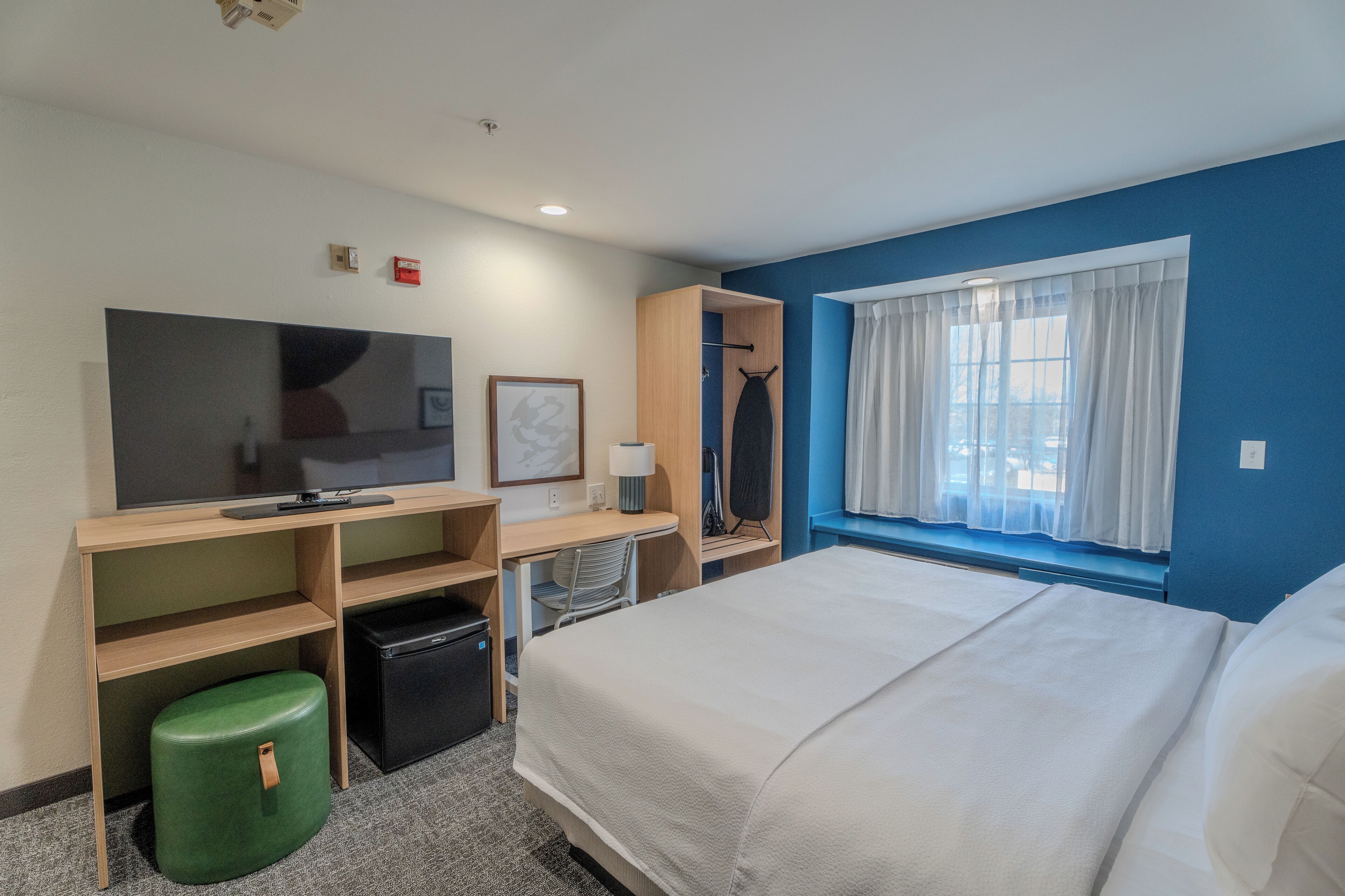 A hotel room with a bed, a desk with a computer, a television, a small green stool, and a window with curtains.