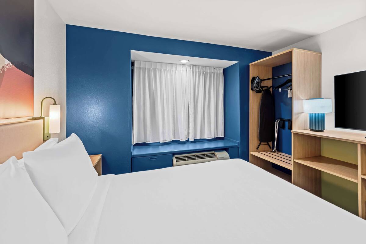 Ein Hotelzimmer mit Bett, Fenster mit weißen Vorhängen, Schrank, Fernseher und Nachttischlampe.