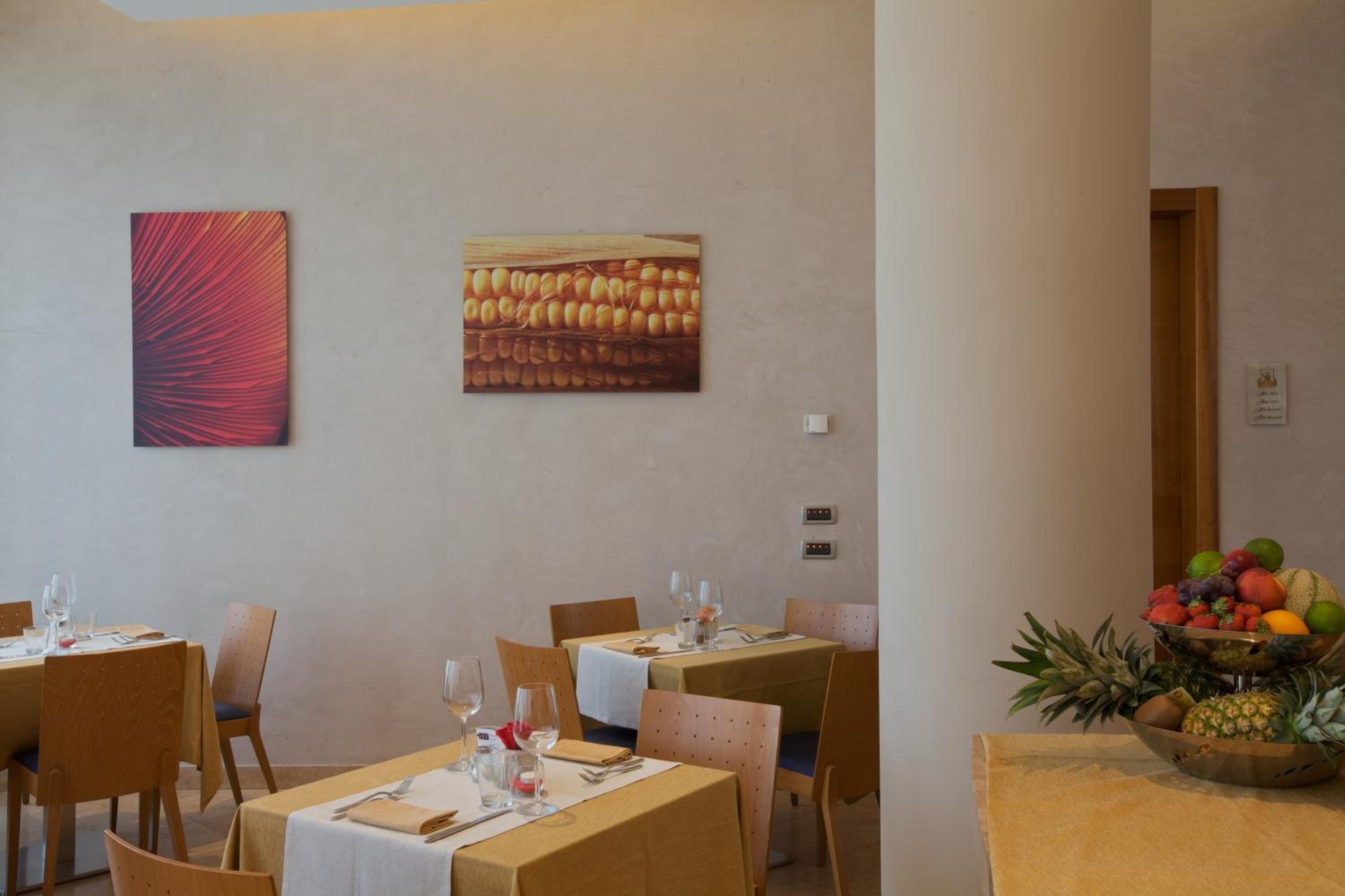 A dining area with tables set for four, each with a white tablecloth, wooden chairs, and a fruit bowl on the right.