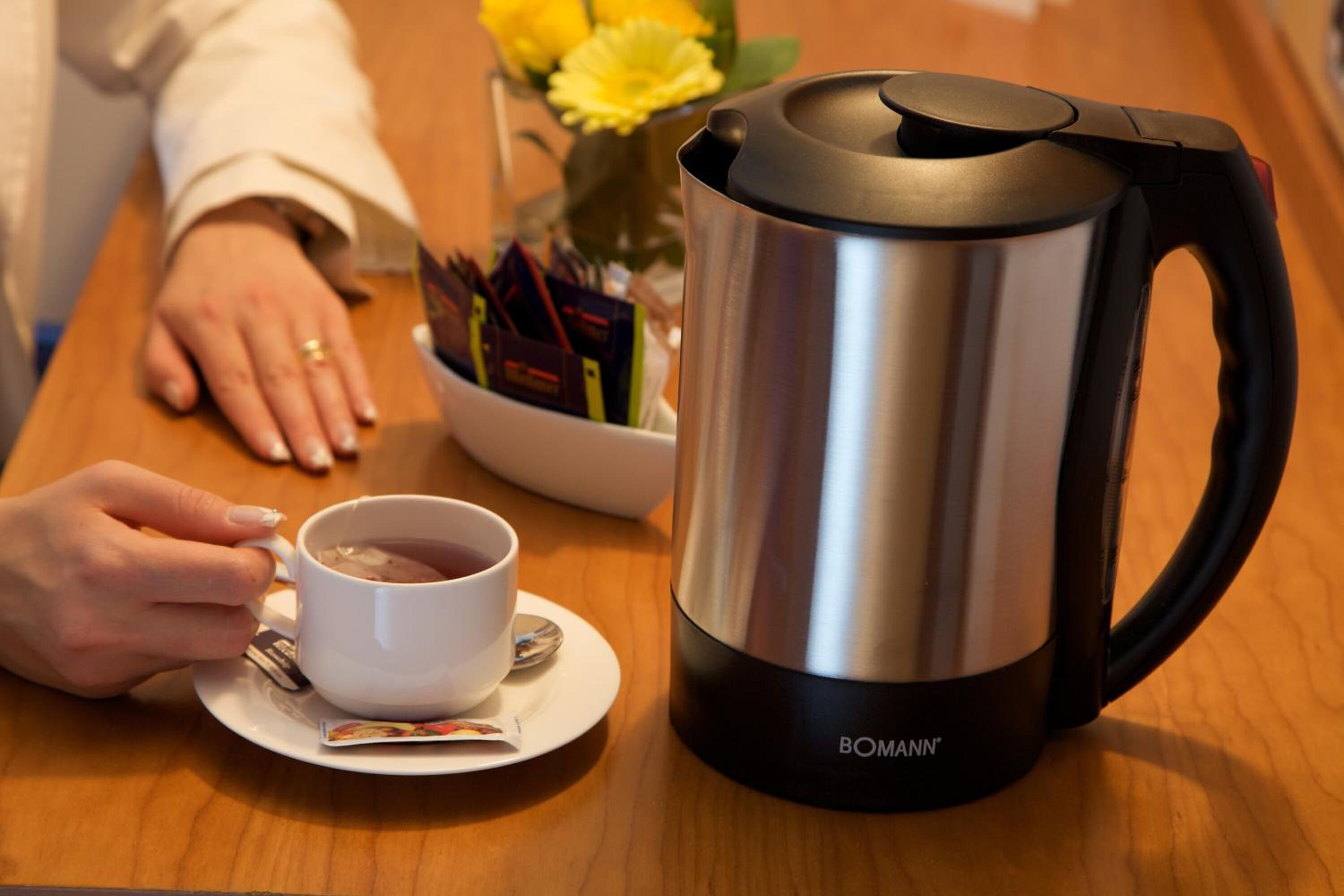 A stainless steel coffee pot with a black handle and a white cup with a saucer on a wooden table.