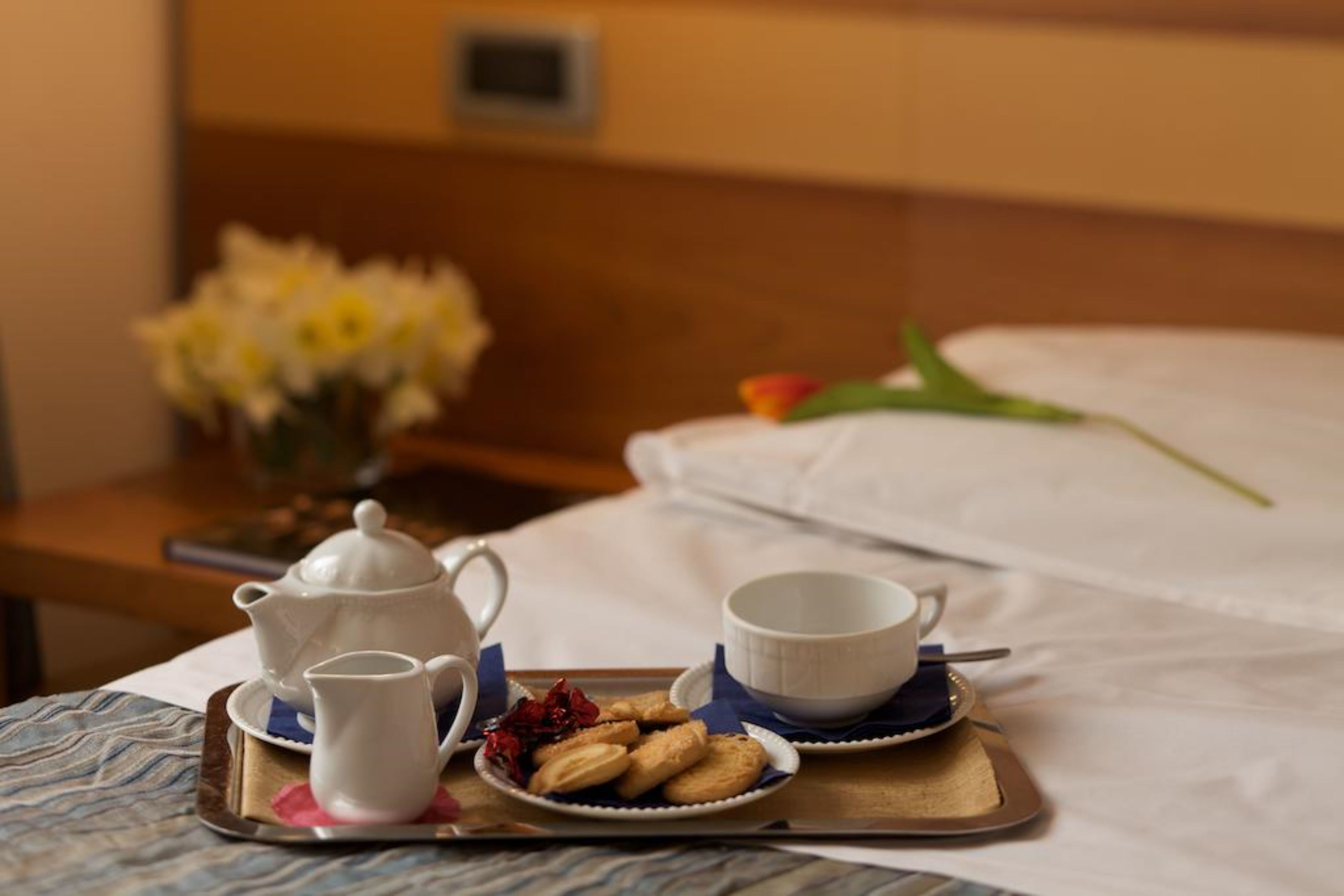 A neatly arranged breakfast tray with a teapot, cups, and cookies on a bed with a white sheet and a vase of flowers in the background.