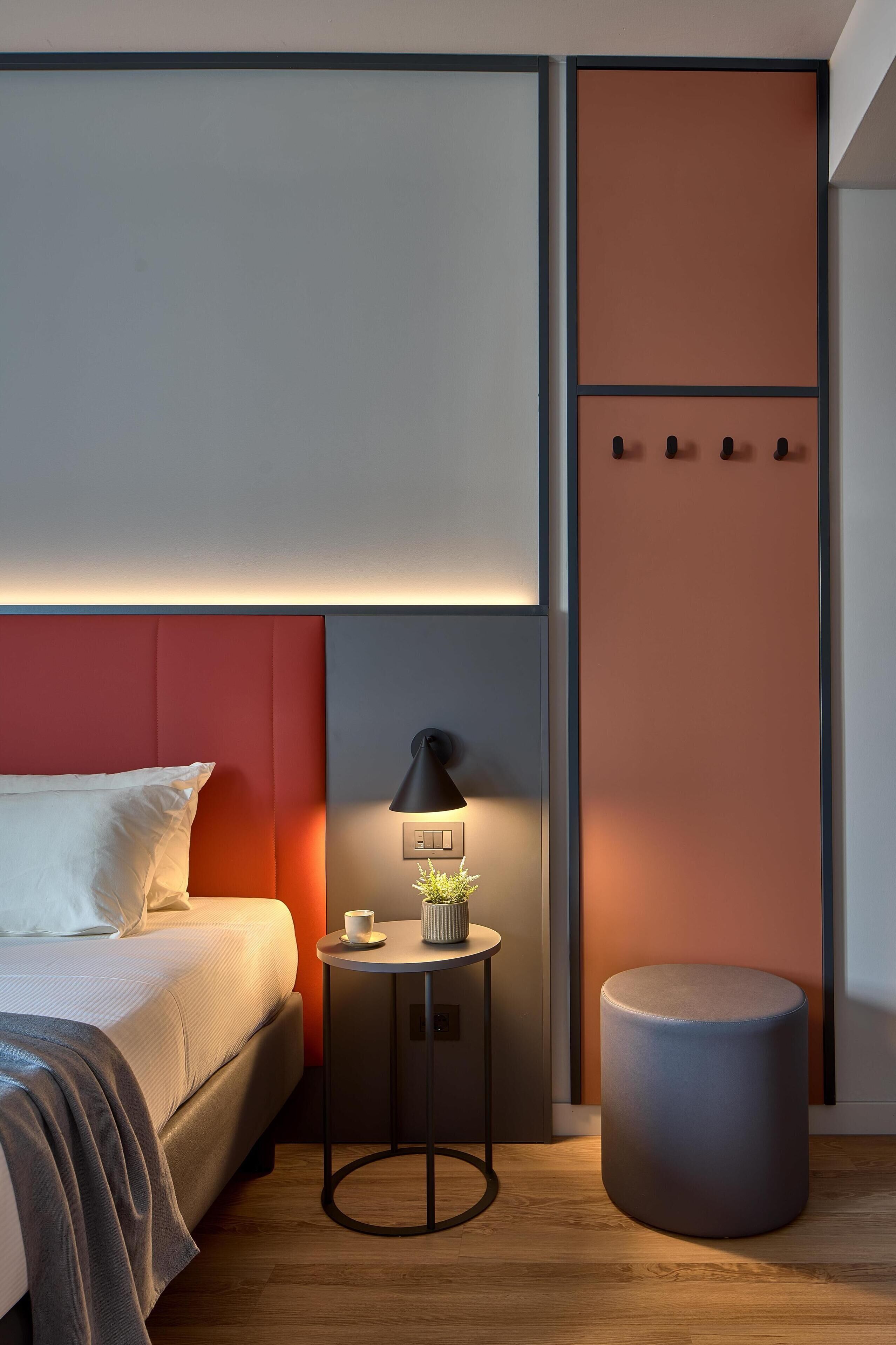 Una moderna camera d'albergo con un letto, un comodino, una lampada e un poggiapiedi rotondo.