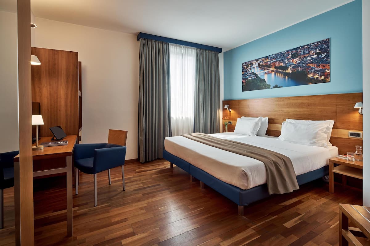 A hotel room with a bed, desk, chair, and a cityscape painting on the wall.