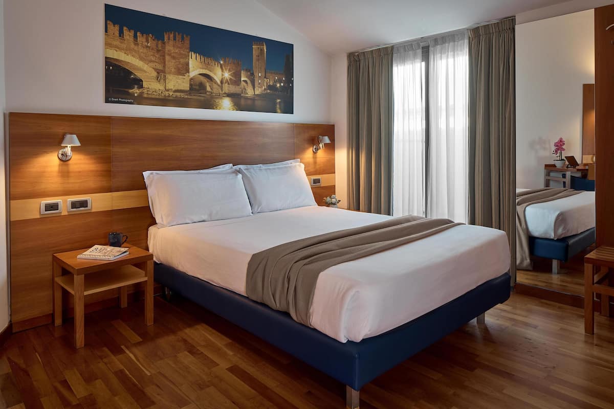 A hotel room with a large bed, wooden headboard, two bedside tables, a nightstand, a desk with a chair, and a large window with curtains.