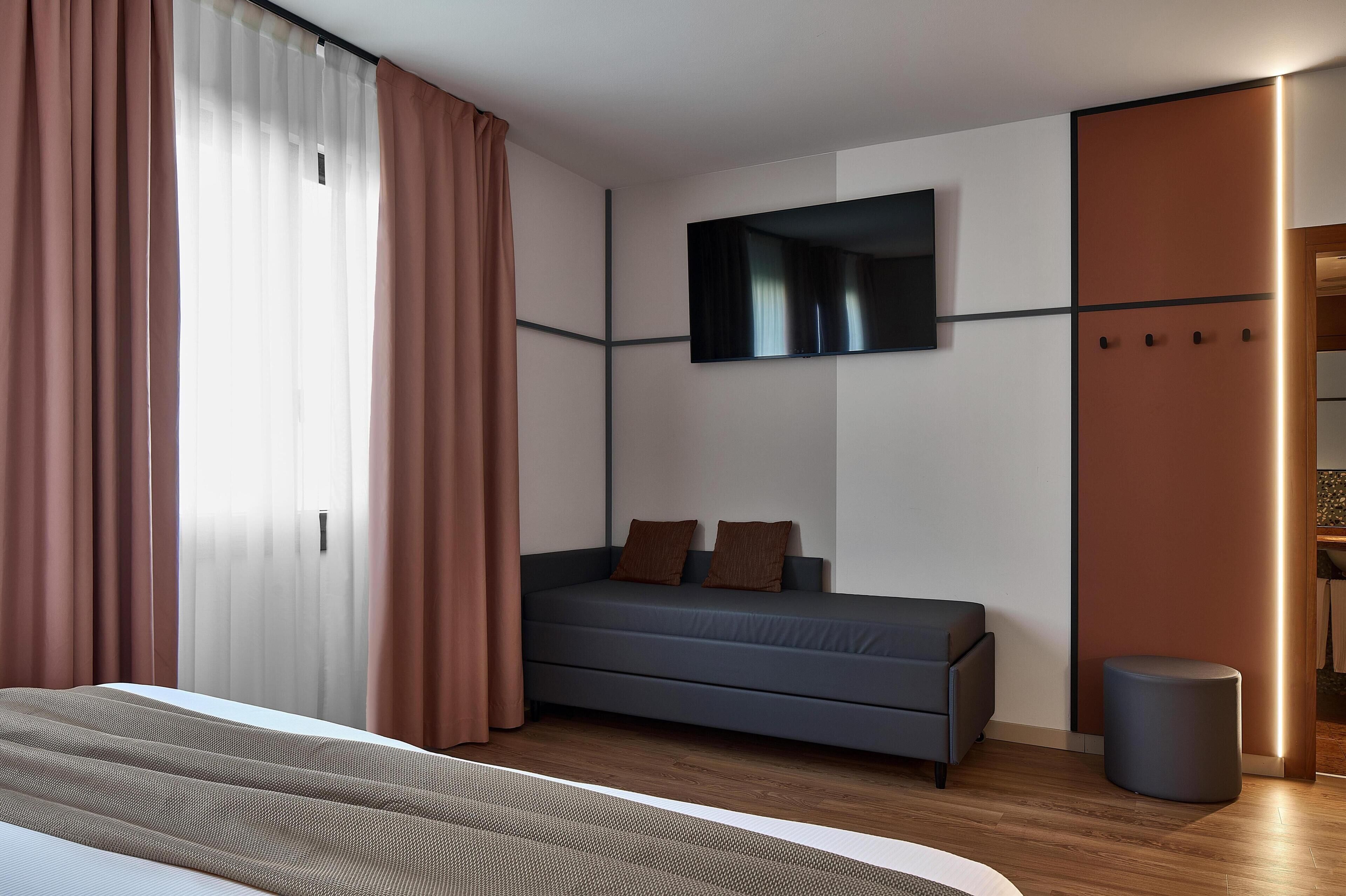 Habitación de hotel moderna con cama, sofá, televisión montada en la pared y un taburete pequeño y redondo.