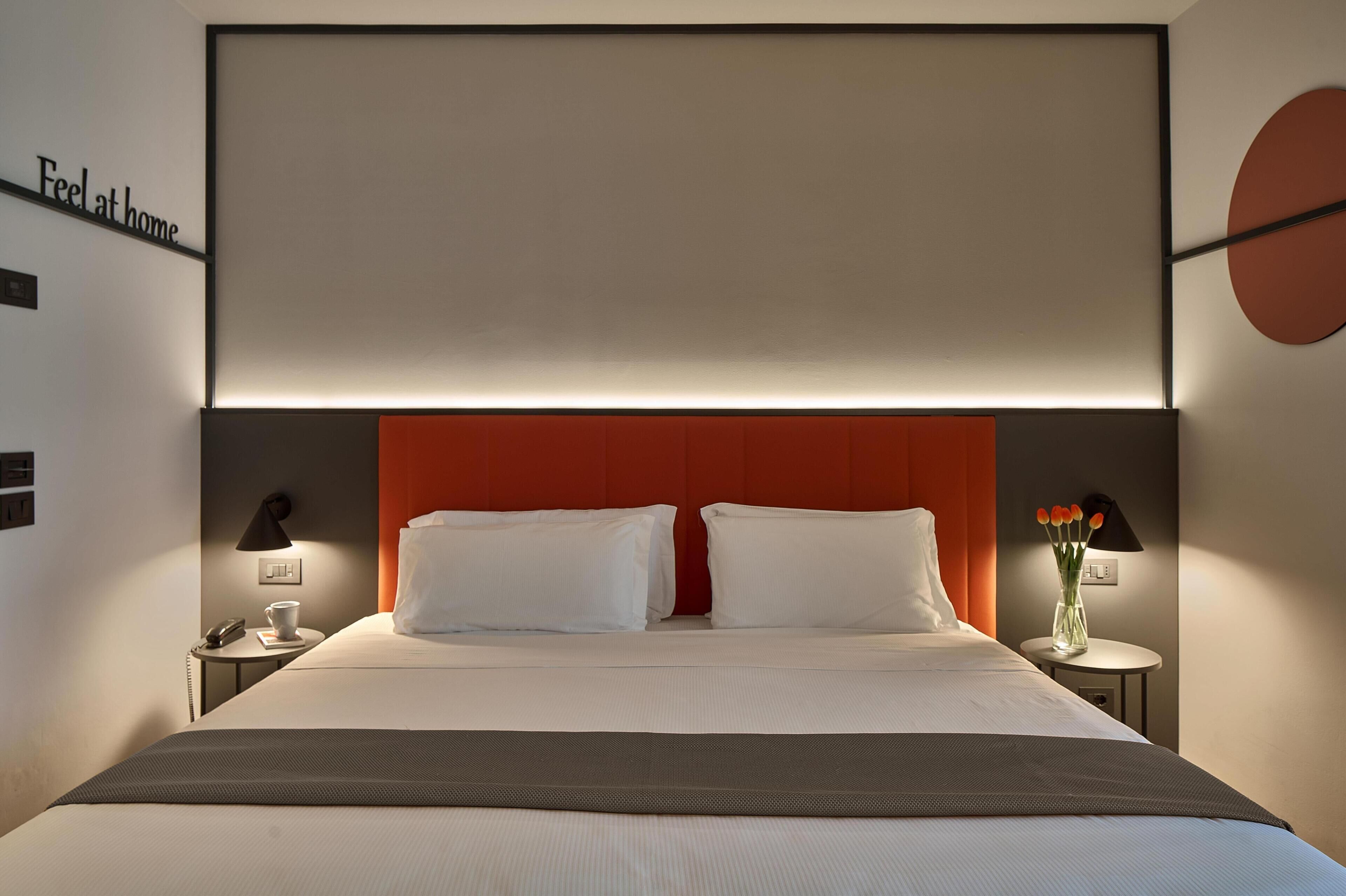 Habitación de hotel con una cama, dos mesitas de noche, una lámpara de pared y un florero con flores.