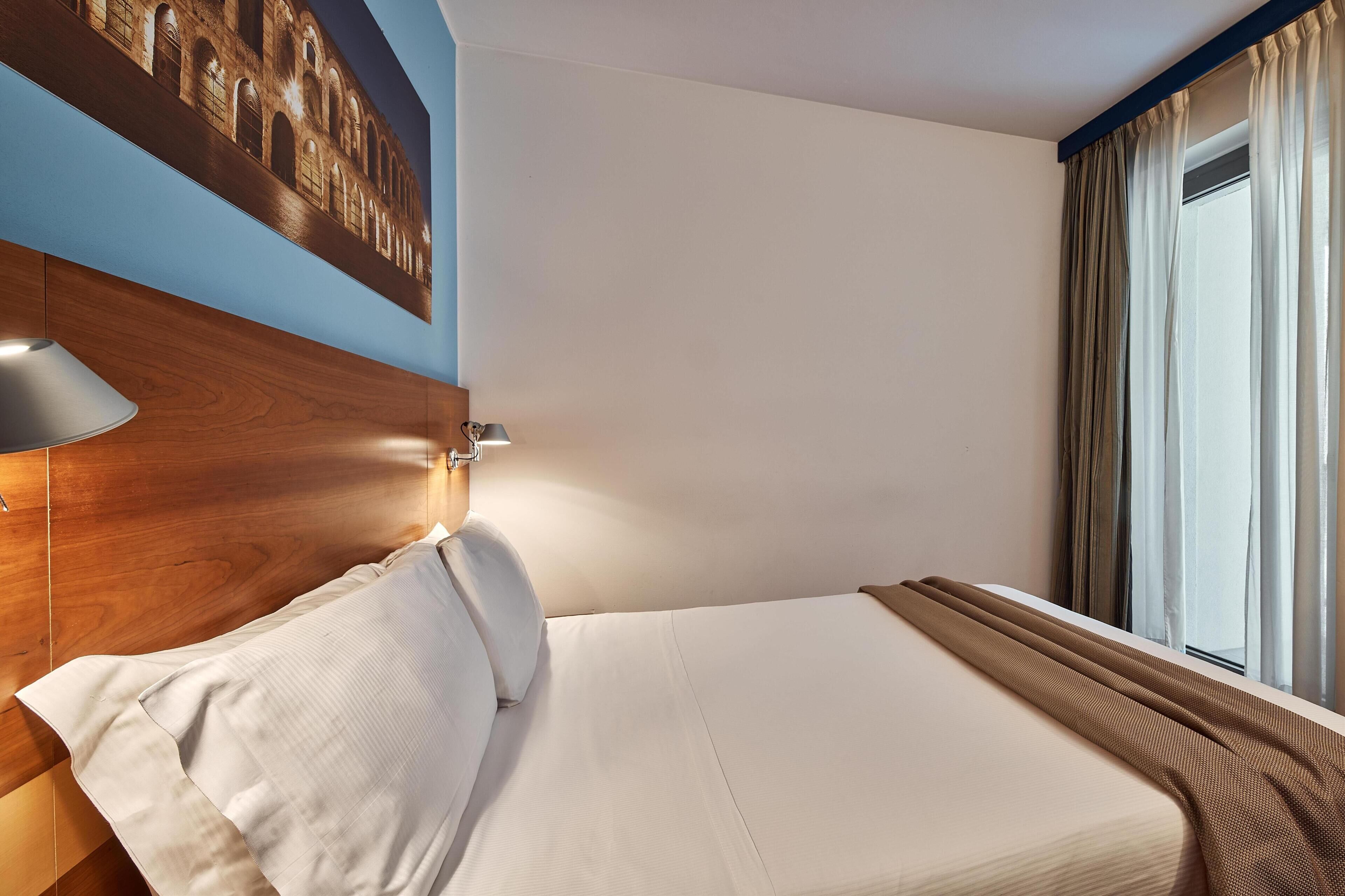 A hotel room with a bed, a wall-mounted lamp, a window with curtains, and a wall art piece.