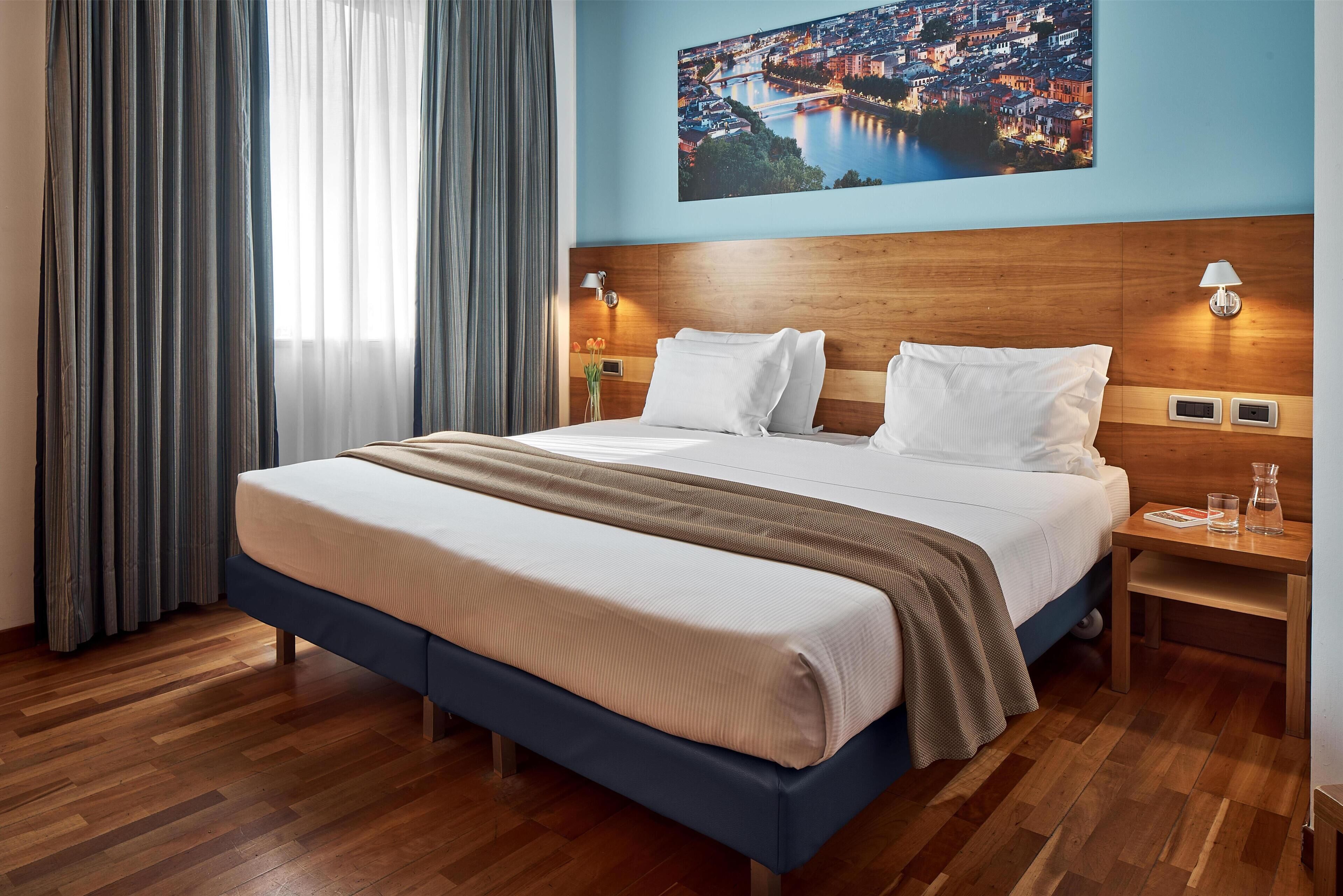 A hotel room with a large bed, wooden headboard, two bedside lamps, a nightstand, and a cityscape view.