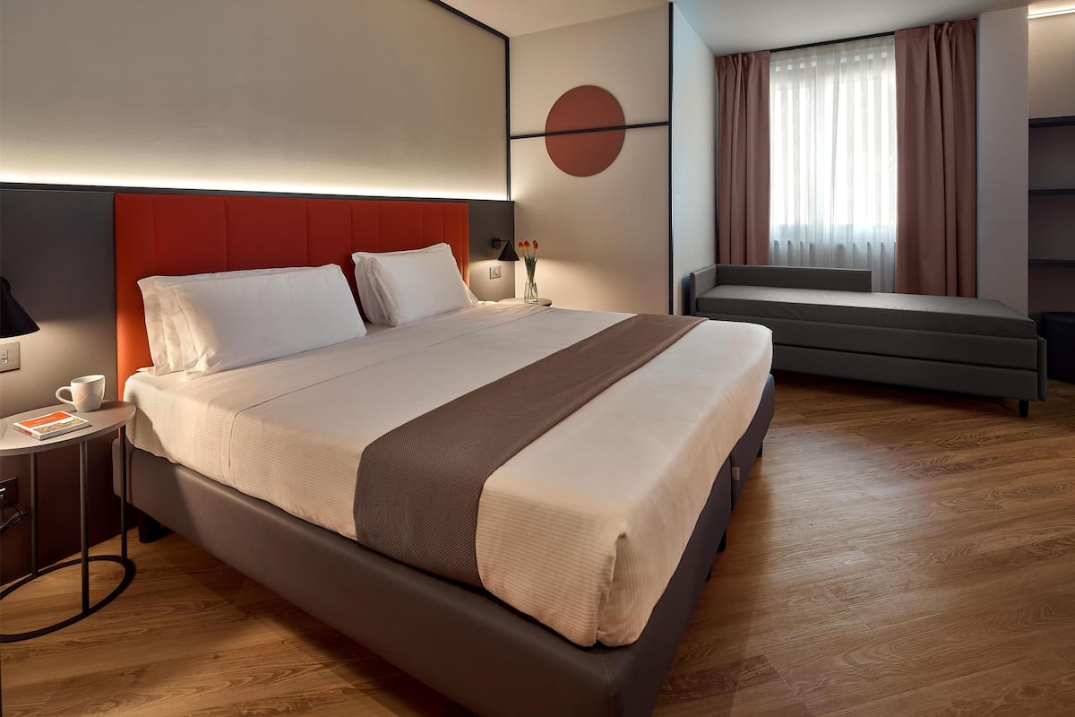 A modern hotel room with a large bed, bedside table, lamp, and a view of the city.
