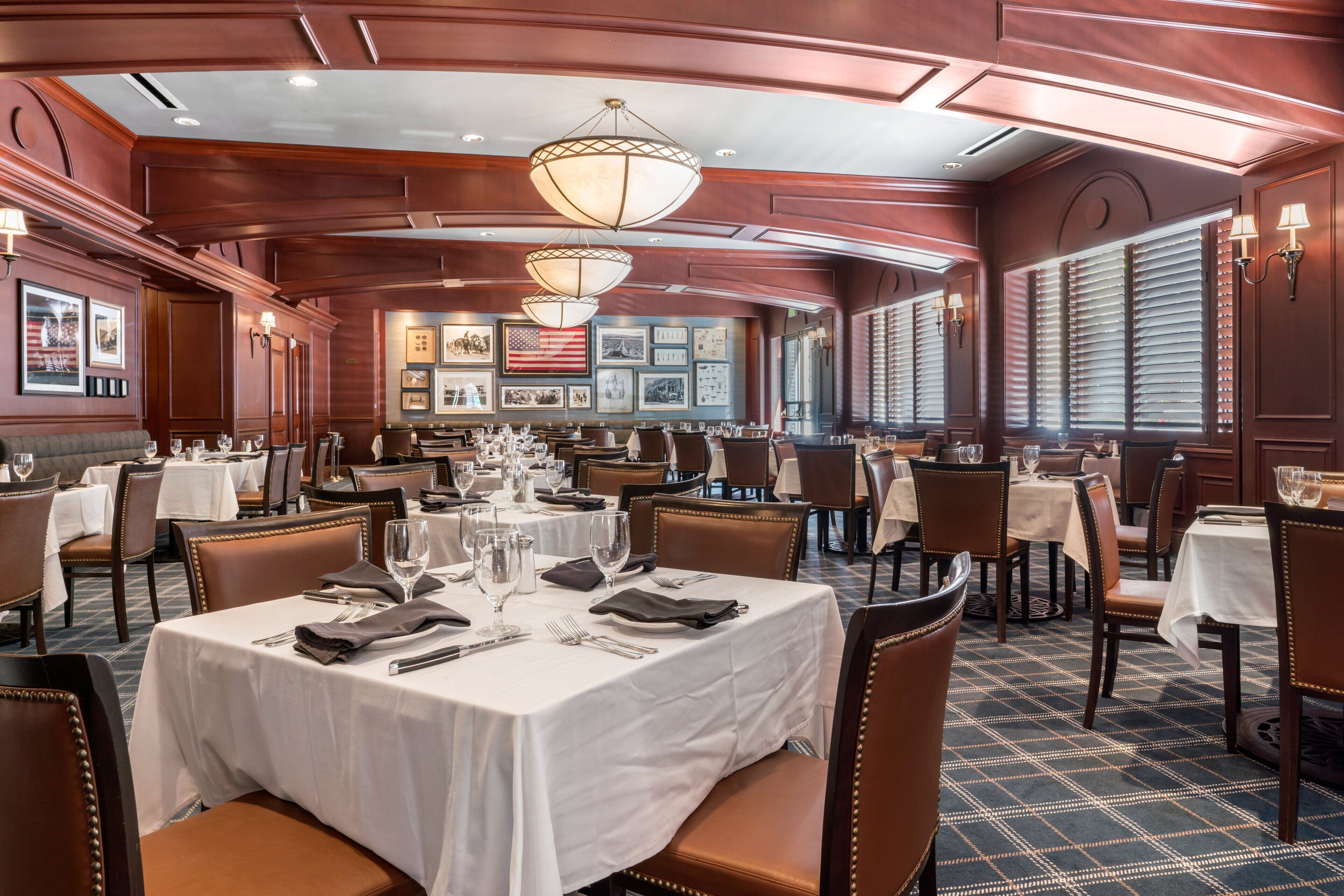 A formal dining area with multiple tables set for service, featuring a classic design with wooden paneling and chandeliers.