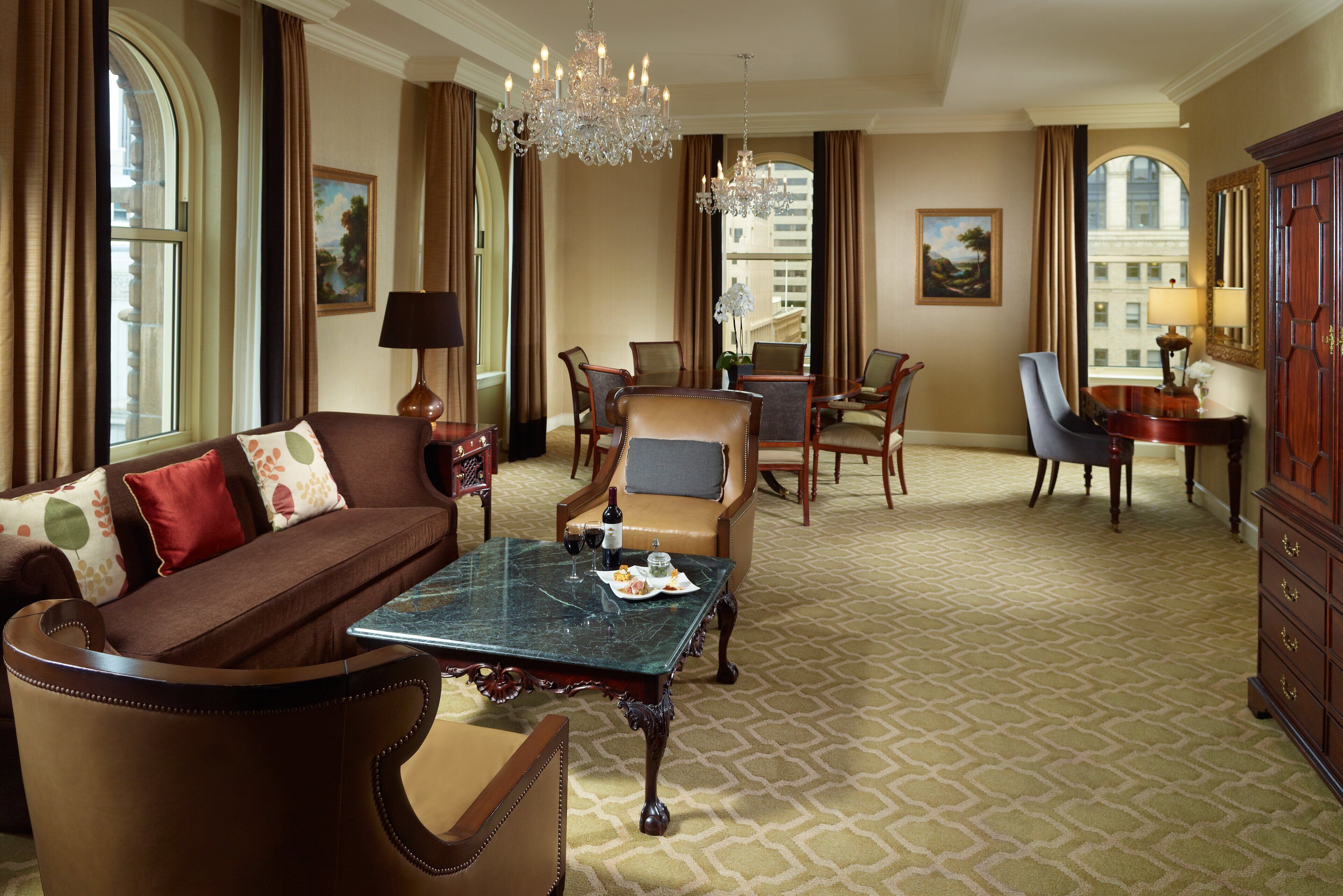 A spacious living room with a brown sofa, a marble coffee table, and a chandelier. There is a dining area with a table and chairs, and a large window with curtains.