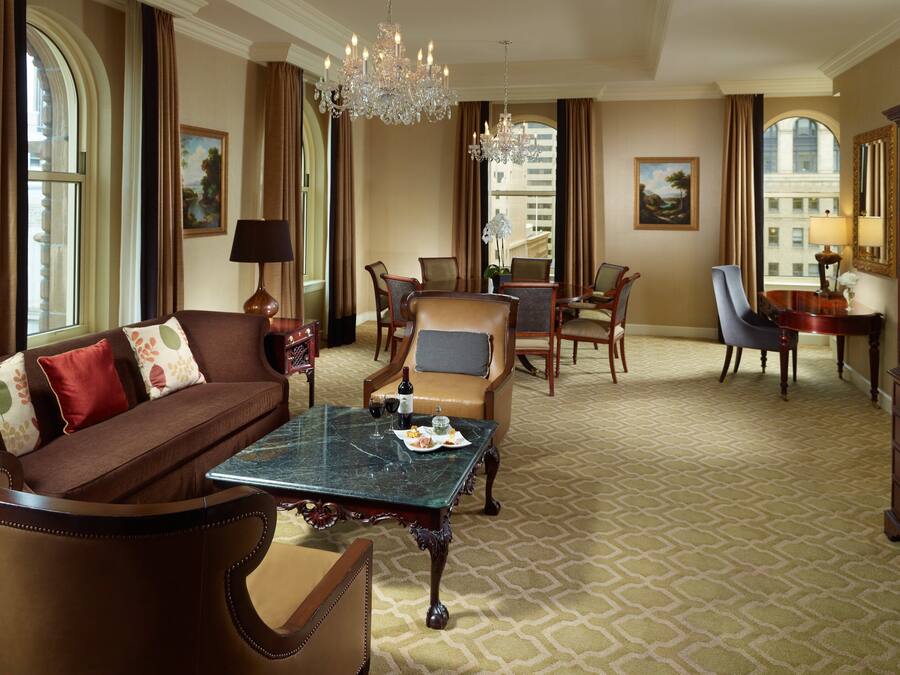 A spacious living room with a brown sofa, a marble coffee table, and a chandelier. There is a dining area with a table and chairs, and a large window with curtains.